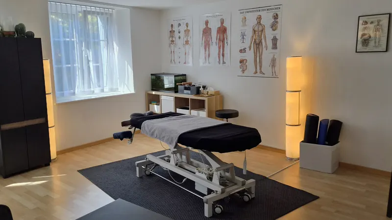 Behandlungsraum mit elektrischer Behandlungsliege, Stehlampe und anatomischen Tafeln